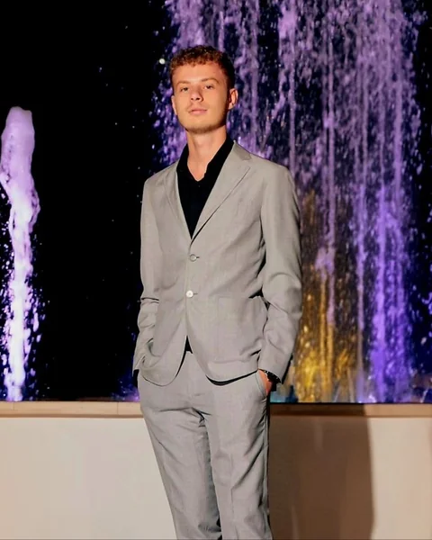 Gabriele Decandia in a grey suit in front of a lit fountain at night